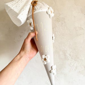May include: A beige cone-shaped school cone decorated with white crocheted flowers and a brown drawstring. The fabric features the name Mali and the date 01.08.20 in gold lettering. A white muslin fabric top with gold dots extends from the top.