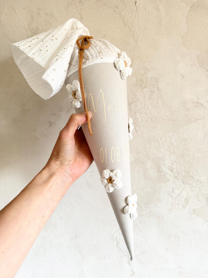 May include: A beige cone-shaped school cone decorated with white crocheted flowers and a brown drawstring. The fabric features the name Mali and the date 01.08.20 in gold lettering. A white muslin fabric top with gold dots extends from the top.