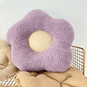 May include: A plush purple flower-shaped cushion with a cream-coloured centre. The cushion is sitting on a tan cushion.