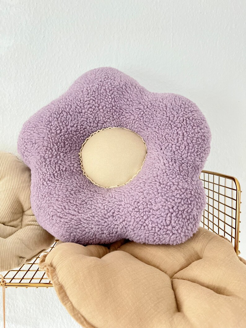 May include: A plush purple flower-shaped cushion with a cream-coloured centre. The cushion is sitting on a tan cushion.