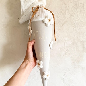 May include: A cone-shaped fabric gift bag in light gray, featuring the name Mali and the date 01.08.20 in gold lettering. The bag is adorned with white crocheted flowers and topped with a white muslin fabric closure tied with a brown cord.