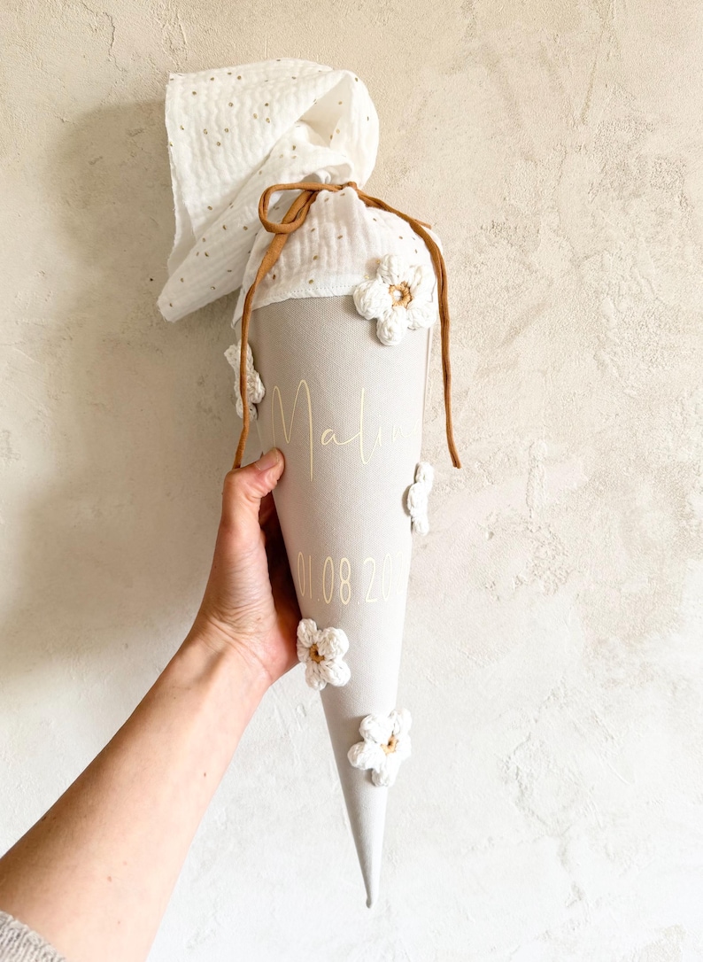 May include: A cone-shaped fabric gift bag in light gray, featuring the name Mali and the date 01.08.20 in gold lettering. The bag is adorned with white crocheted flowers and topped with a white muslin fabric closure tied with a brown cord.