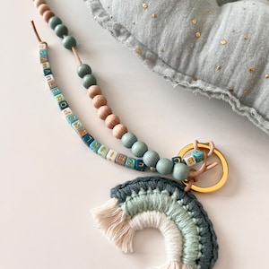Puede incluir: Un llavero de macramé arcoíris azul y blanco con una cadena de cuentas de madera y cuentas de letras. El llavero está hecho con hilo azul claro, blanco y azul oscuro.