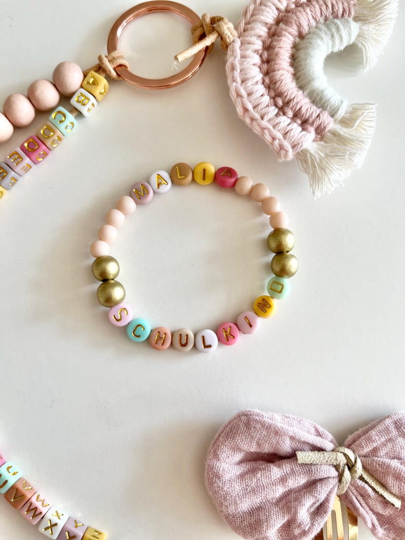 Pu&ograve; includere: Un braccialetto di perline con il nome "MALIA SCHULKIND" scritto con perline di lettere colorate. Il braccialetto presenta perline dorate e rosa, insieme a un portachiavi in oro rosa, un arcobaleno in macram&egrave; e un fiocco rosa.
