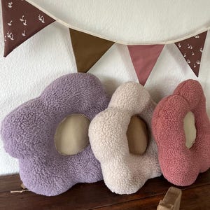 May include: Three plush flower-shaped cushions in purple, cream, and pink. The cushions have a hole in the centre and are made of soft, fuzzy fabric.