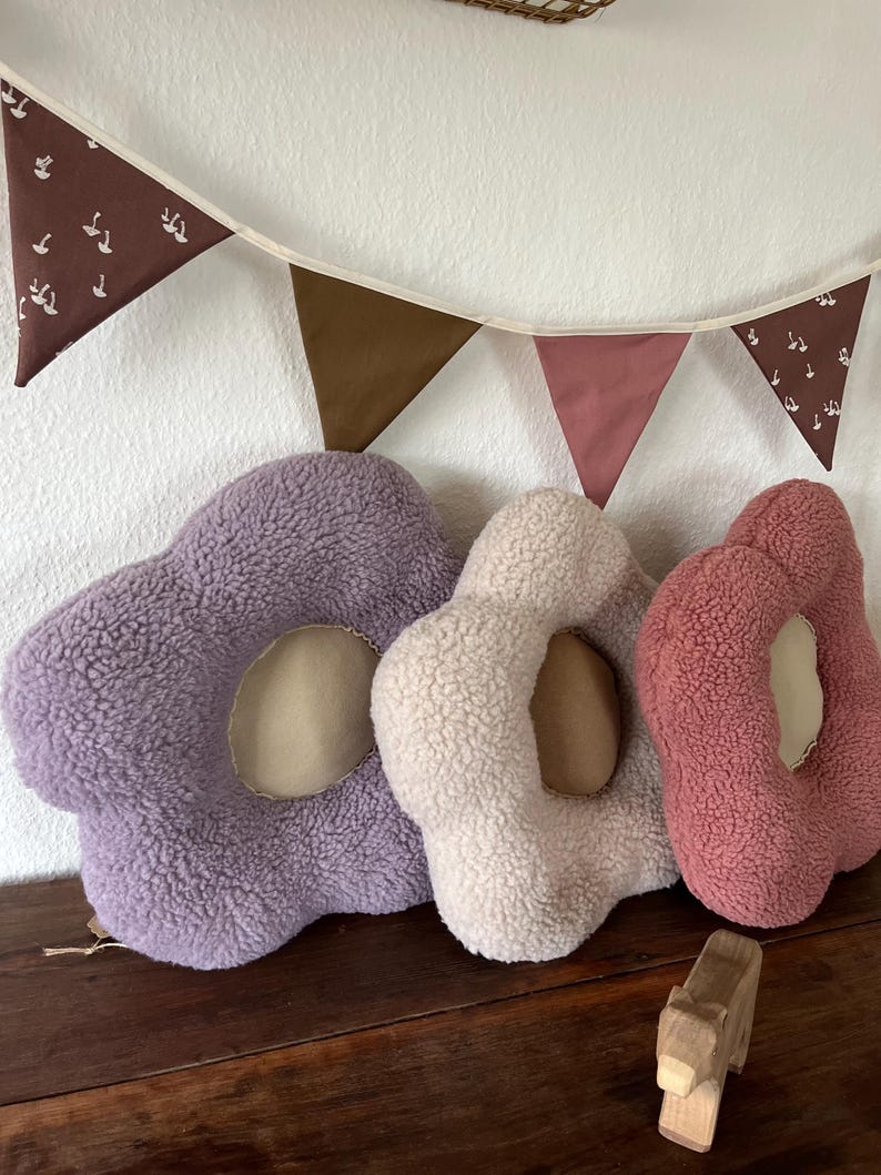 May include: Three plush flower-shaped cushions in purple, cream, and pink. The cushions have a hole in the centre and are made of soft, fuzzy fabric.
