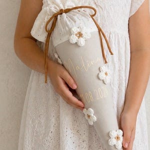 May include: A beige fabric cone-shaped gift bag featuring white crocheted flower appliqu&eacute;s, a white eyelet fabric top, and brown cord drawstring ties. The bag is personalized with the name Malina and the date 11.08.2024 in gold cursive lettering.