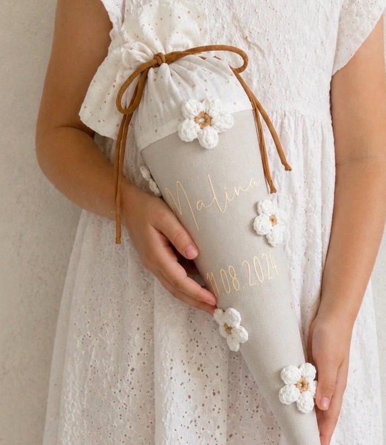 May include: A beige fabric cone-shaped gift bag featuring white crocheted flower appliqu&eacute;s, a white eyelet fabric top, and brown cord drawstring ties. The bag is personalized with the name Malina and the date 11.08.2024 in gold cursive lettering.