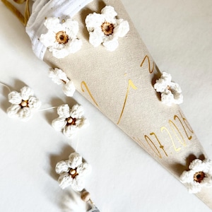May include: A beige fabric cone decorated with white crocheted flowers, gold lettering, and a white muslin top tied with a gold ribbon. A string of white crocheted flowers with a clear crystal pendant hangs from the side of the cone.