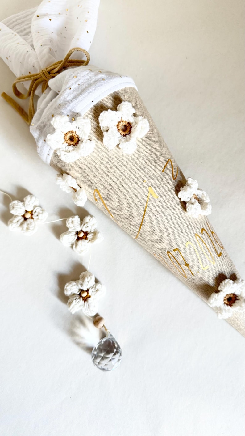 May include: A beige fabric cone decorated with white crocheted flowers, gold lettering, and a white muslin top tied with a gold ribbon. A string of white crocheted flowers with a clear crystal pendant hangs from the side of the cone.