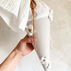 May include: A beige cone-shaped gift bag featuring white crocheted flowers, a white fabric top with gold polka dots, and a brown drawstring. The bag is personalized with the name "Mali" and the date "01.08" printed in a gold script font.