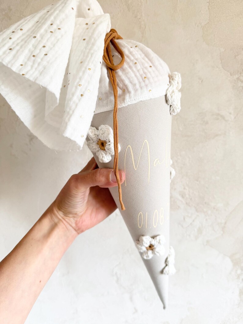 May include: A beige cone-shaped gift bag featuring white crocheted flowers, a white fabric top with gold polka dots, and a brown drawstring. The bag is personalized with the name "Mali" and the date "01.08" printed in a gold script font.