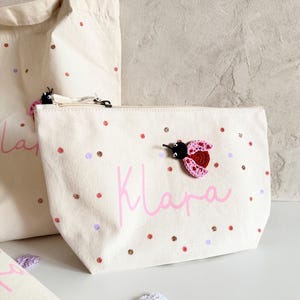 Op de afbeelding: Een crèmekleurig canvas tasje met ritssluiting voorzien van de naam Klara in roze schrijfletters, versierd met verspreide roze en paarse stippen en een kleine, driedimensionale gehaakte lieveheersbeestje-applicatie met roze vleugels.