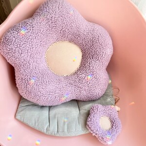 May include: A plush lavender flower-shaped cushion with a smaller matching cushion. The cushions are sitting on a light pink chair with a grey corduroy cushion.