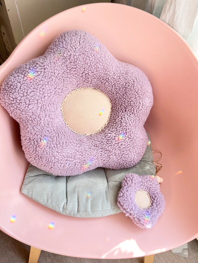May include: A plush lavender flower-shaped cushion with a smaller matching cushion. The cushions are sitting on a light pink chair with a grey corduroy cushion.
