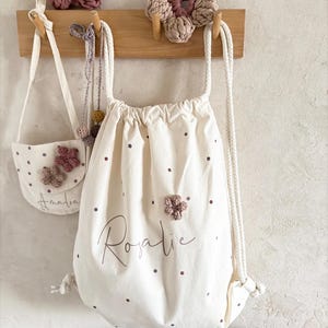 Könnte beinhalten: Ein cremefarbener Kordelzug-Rucksack mit dunklen Punkten und dem Namen Rosalie in Schreibschrift. Eine kleinere passende Tasche mit dem Namen Amalia ist daneben. Beide Taschen haben kleine gehäkelte Blumenapplikationen in Altrosa und Beige.