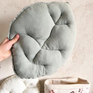 Pode incluir: Uma almofada em forma de folha verde claro com uma superfície texturizada é segurada por uma mão. Abaixo, uma almofada em forma de folha branca e uma cesta de tecido bege com enfeites florais são visíveis.
