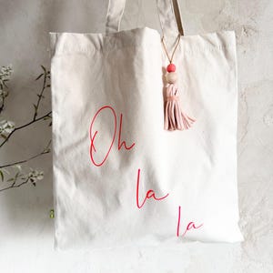 Op de afbeelding: Crèmekleurige draagtas met rode tekst "Oh la la". De tas heeft twee handvatten en een decoratieve kwast met een roze kwast en houten kralen. Een stijlvol en praktisch accessoire.