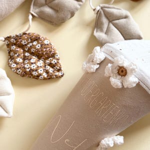 May include: A beige fabric cone featuring the text "KINDERGARTENKIND" and a name, decorated with white crocheted flowers. A garland of fabric leaves in shades of brown, beige, and a floral print is positioned above the cone on a light surface.