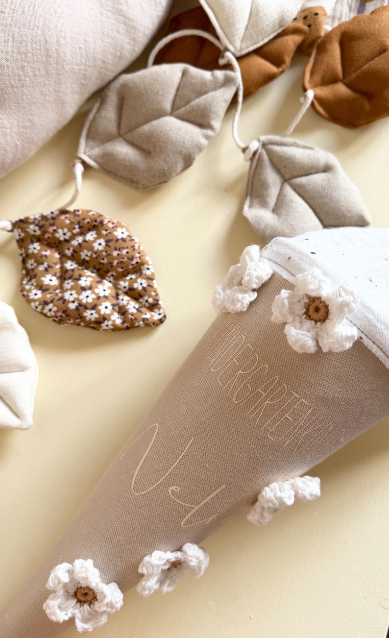 May include: A beige fabric cone featuring the text "KINDERGARTENKIND" and a name, decorated with white crocheted flowers. A garland of fabric leaves in shades of brown, beige, and a floral print is positioned above the cone on a light surface.