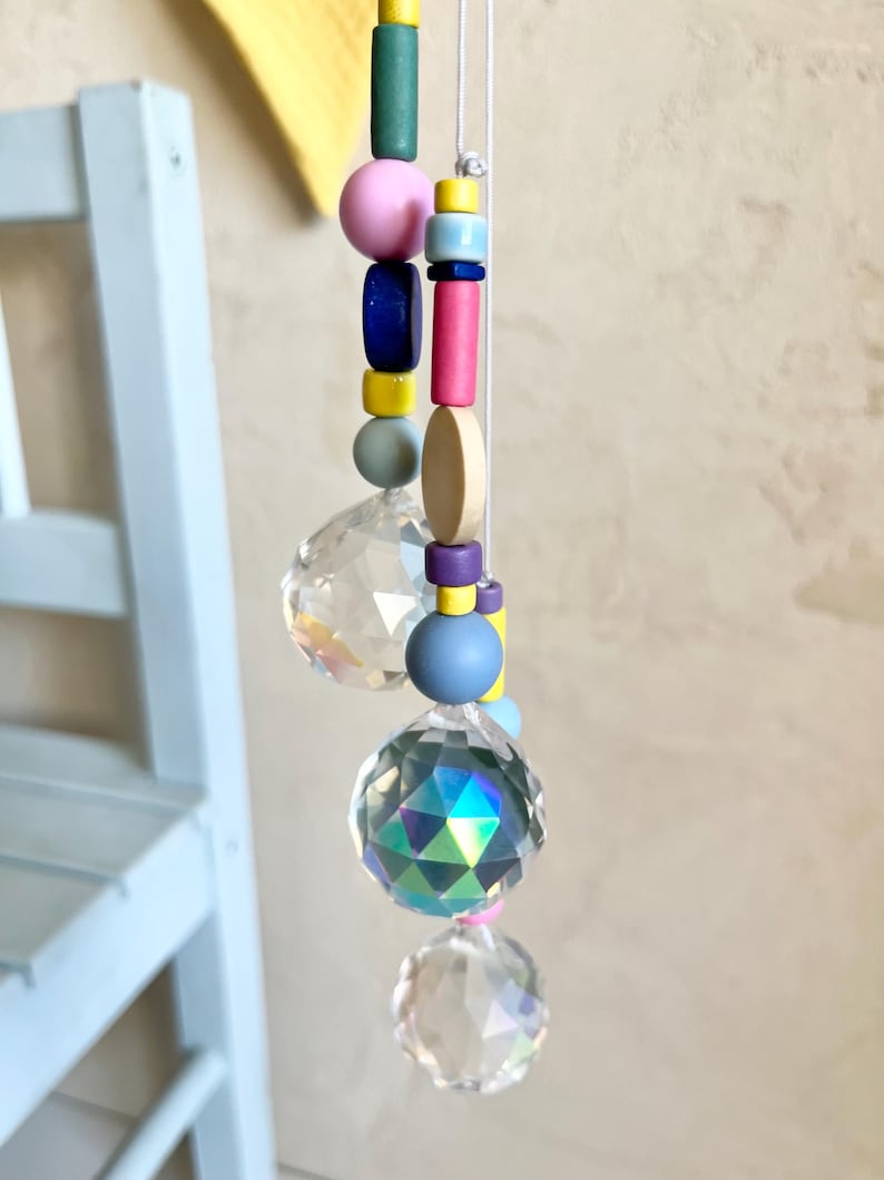 Peut inclure: Un attrape-soleil en cristal avec trois prismes en cristal transparent et une cha&icirc;ne de perles avec des perles en bois color&eacute;es dans les tons rose, bleu, vert, jaune et violet.