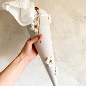 May include: A light gray conical gift bag featuring white crocheted flower appliqu&eacute;s with brown centers. The top is finished with a white eyelet fabric closure secured by a thin brown cord. The surface displays faint white text printed vertically.