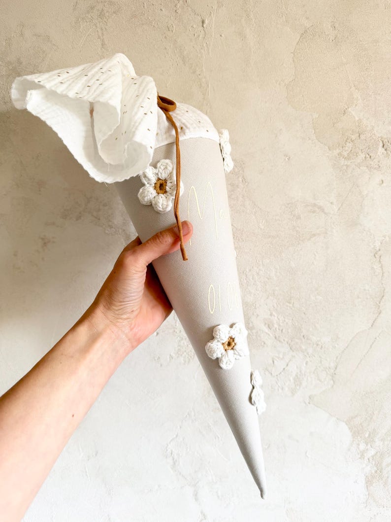 May include: A light gray conical gift bag featuring white crocheted flower appliqu&eacute;s with brown centers. The top is finished with a white eyelet fabric closure secured by a thin brown cord. The surface displays faint white text printed vertically.