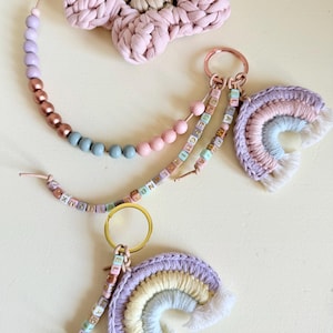 Puede incluir: Una colección de llaveros y accesorios hechos a mano. La imagen presenta una gran flor de ganchillo rosa, llaveros de cuentas con colores pastel y dos llaveros de arcoíris de ganchillo. Los llaveros tienen herrajes dorados y oro rosa.