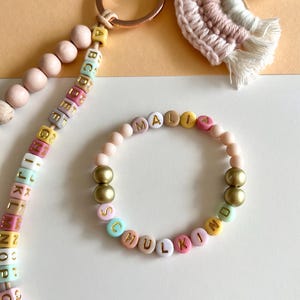 Pu&ograve; includere: Un braccialetto e un portachiavi personalizzati con perline colorate con le lettere che compongono le parole "MALIA" e "SCHULKIND". Il braccialetto presenta perline color oro. Sono visibili anche un portachiavi con perline di legno e un arcobaleno in macram&egrave;.