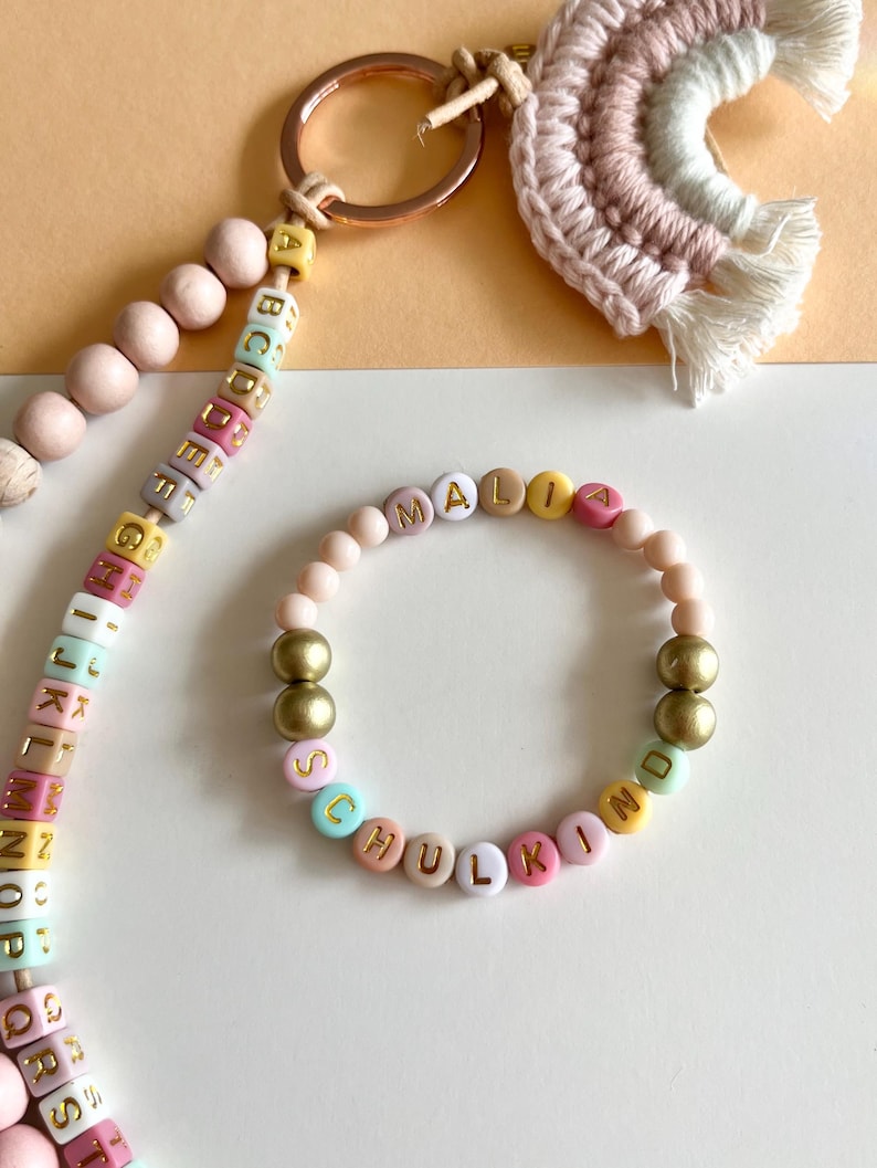Pu&ograve; includere: Un braccialetto e un portachiavi personalizzati con perline colorate con le lettere che compongono le parole "MALIA" e "SCHULKIND". Il braccialetto presenta perline color oro. Sono visibili anche un portachiavi con perline di legno e un arcobaleno in macram&egrave;.