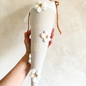 May include: A beige cone-shaped gift bag featuring white crocheted flower appliqu&eacute;s, a white fabric top with brown drawstring ties, and the gold text "Alina" and "2026" printed on the front surface of the fabric cone.