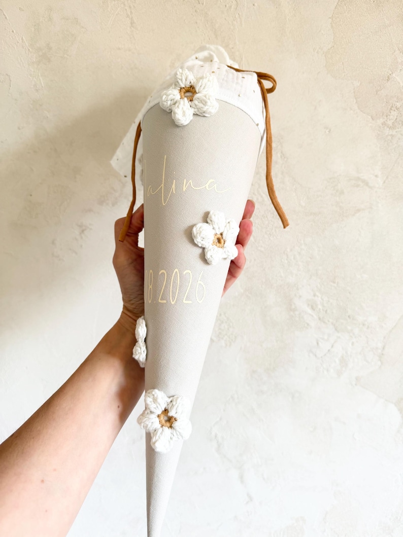 May include: A beige cone-shaped gift bag featuring white crocheted flower appliqu&eacute;s, a white fabric top with brown drawstring ties, and the gold text "Alina" and "2026" printed on the front surface of the fabric cone.