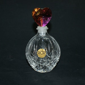 May include: A clear glass bottle with a heart-shaped stopper. The stopper is iridescent and has shades of pink, orange, and yellow. The bottle has a label that reads "RCR, Royal Crystal Rock, Dalia".
