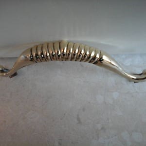 May include: A gold-toned metal cabinet door handle with a curved, ribbed design.