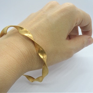 Puede incluir: Un brazalete de oro retorcido. El brazalete está hecho de un metal grueso y texturizado y tiene un diseño simple y elegante.