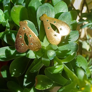 Peut inclure: Une paire de boucles d'oreilles dorées de forme originale, ornées de petites pierres vertes. Les boucles d'oreilles sont présentées sur un fond de feuilles succulentes vertes et de fleurs jaunes, créant une esthétique naturelle.