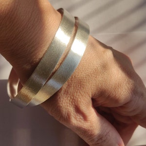 Puede incluir: Dos pulseras de plata apiladas en la muñeca de una persona. Ambas pulseras son lisas y brillantes.