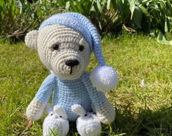 May include: A white crocheted teddy bear wearing a blue crocheted sleep hat and holding two white crocheted bunny slippers. The bear is sitting on a bed of green grass.