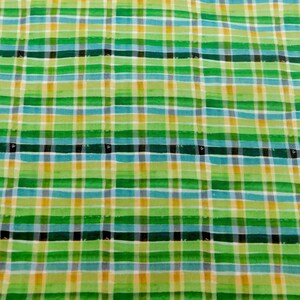 May include: Close-up of a fabric with a plaid pattern. The design features a grid of intersecting lines in shades of green, blue, yellow, white, and black. The fabric has a textured appearance, suggesting a woven material.