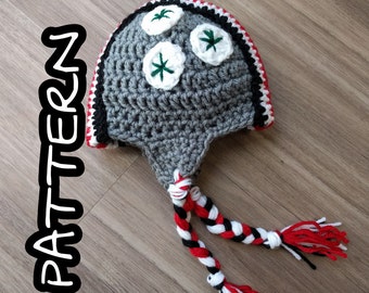 May include: Gray crocheted hat with a white and red trim. The hat has three white crocheted flowers with green centers on top. The hat has two long white, red, and black braided cords.