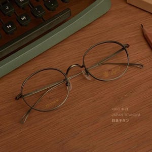 Puede incluir: Gafas redondas con montura oscura y lentes transparentes, etiquetadas como "TITANIUM". Las gafas descansan sobre una superficie de madera, con el texto "KIKO キコ JAPAN TITANIUM 日本チタン."
