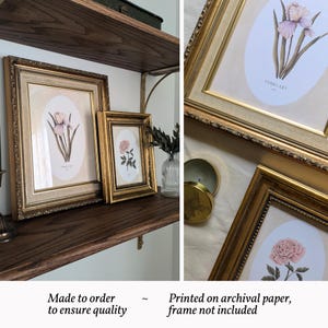May include: Two ornate gold-toned frames containing botanical prints. The larger frame displays an iris illustration labeled FEBRUARY, while the smaller frame features a pink peony illustration. Both prints are set within oval white matting.