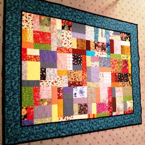 Könnte beinhalten: Ein bunter Patchwork-Deckenbezug mit einem blaugrünen Rand. Die Decke besteht aus vielen verschiedenen Stoffen, darunter Blumenmuster, geometrische Muster und einfarbige Stoffe.