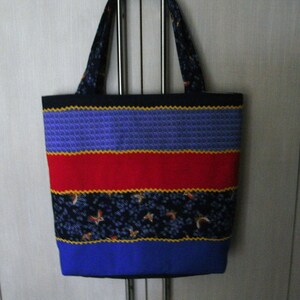 May include: A colorful tote bag with a patchwork design. The bag features a black top, a blue section, a red stripe, and a bottom section with a blue base. The handles and a section of the bag have a dark blue floral pattern.