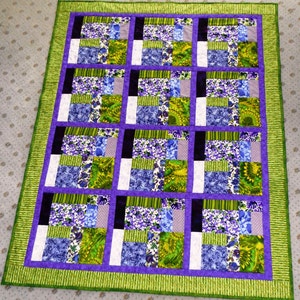 Op de afbeelding: Een paars en groen patchwork-deken met een bloemenpatroon. Het deken is gemaakt van vierkanten van verschillende stoffen, elk met een ander bloemenontwerp. Het deken is afgewerkt met een paarse rand.