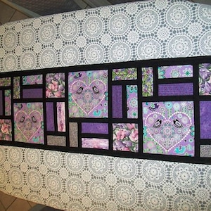 Könnte beinhalten: Ein lila-schwarzer Patchwork-Tischläufer mit geometrischem Muster mit Herzen und Vögeln. Der Läufer besteht aus Stoffquadraten, die mit schwarzem Stoff zusammengenäht sind, der die Kanten umgibt.