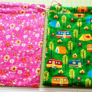 May include: Two drawstring bags, one pink with a floral pattern and one green with a camping themed pattern. Both bags have white drawstrings.