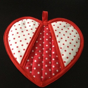 Puede incluir: Un agarrador de olla en forma de corazón rojo y blanco con lunares blancos sobre un fondo rojo. El agarrador de olla tiene un lazo para colgar.
