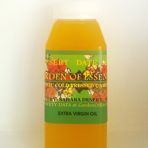 May include: A clear glass bottle with a white cap filled with a yellow liquid. The label is green with a floral design and the text "Desert Date Garden of Essenic Cold Pressed Unrefined Extra Virgin Oil".
