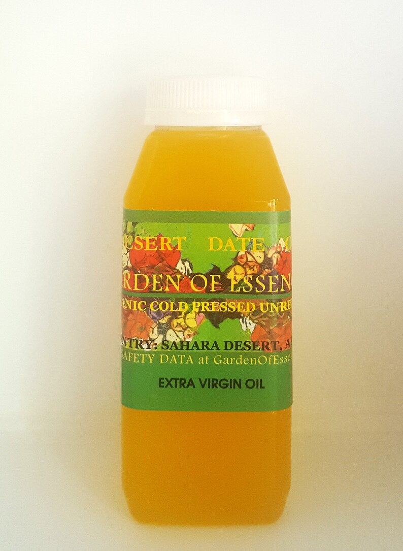 May include: A clear glass bottle with a white cap filled with a yellow liquid. The label is green with a floral design and the text "Desert Date Garden of Essenic Cold Pressed Unrefined Extra Virgin Oil".